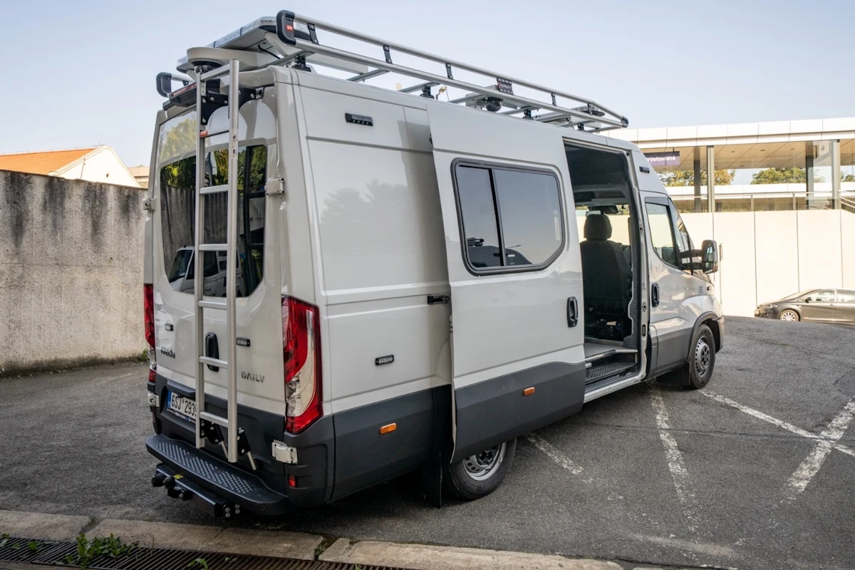 Biały van zaparkowany na placu, jedno z tylnych drzwi jest otwarte, widoczna drabinka prowadząca na dach oraz aluminiowy bagażnik dachowy.