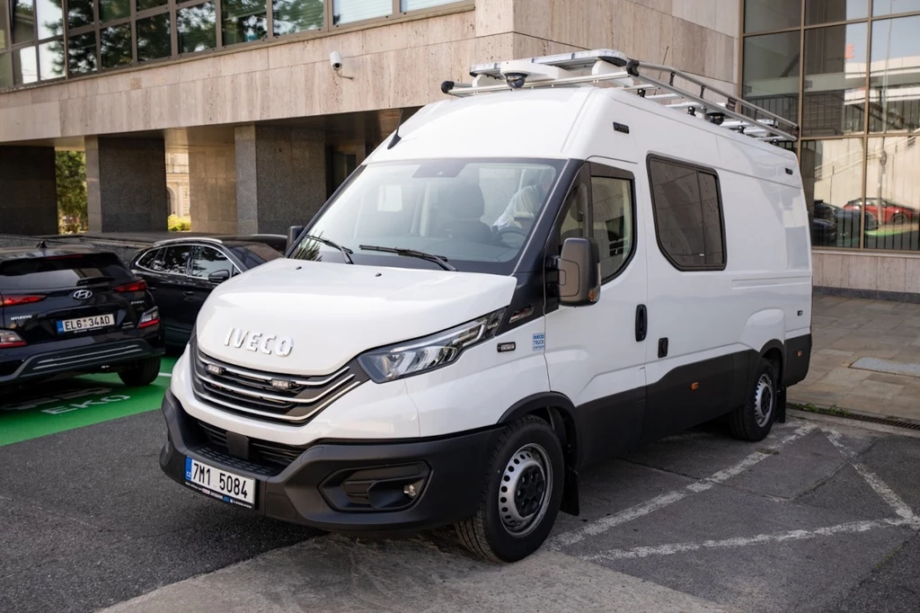 Biały van marki Iveco zaparkowany na ulicy przy nowoczesnym budynku biurowym, z drabiną zamontowaną na dachu oraz liczbową tablicą rejestracyjną z przodu.