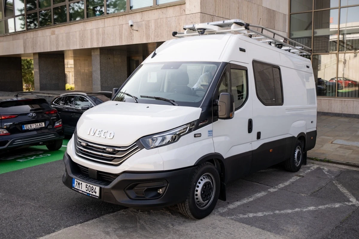 Biały van marki Iveco zaparkowany na ulicy przy nowoczesnym budynku biurowym, z drabiną zamontowaną na dachu oraz liczbową tablicą rejestracyjną z przodu.