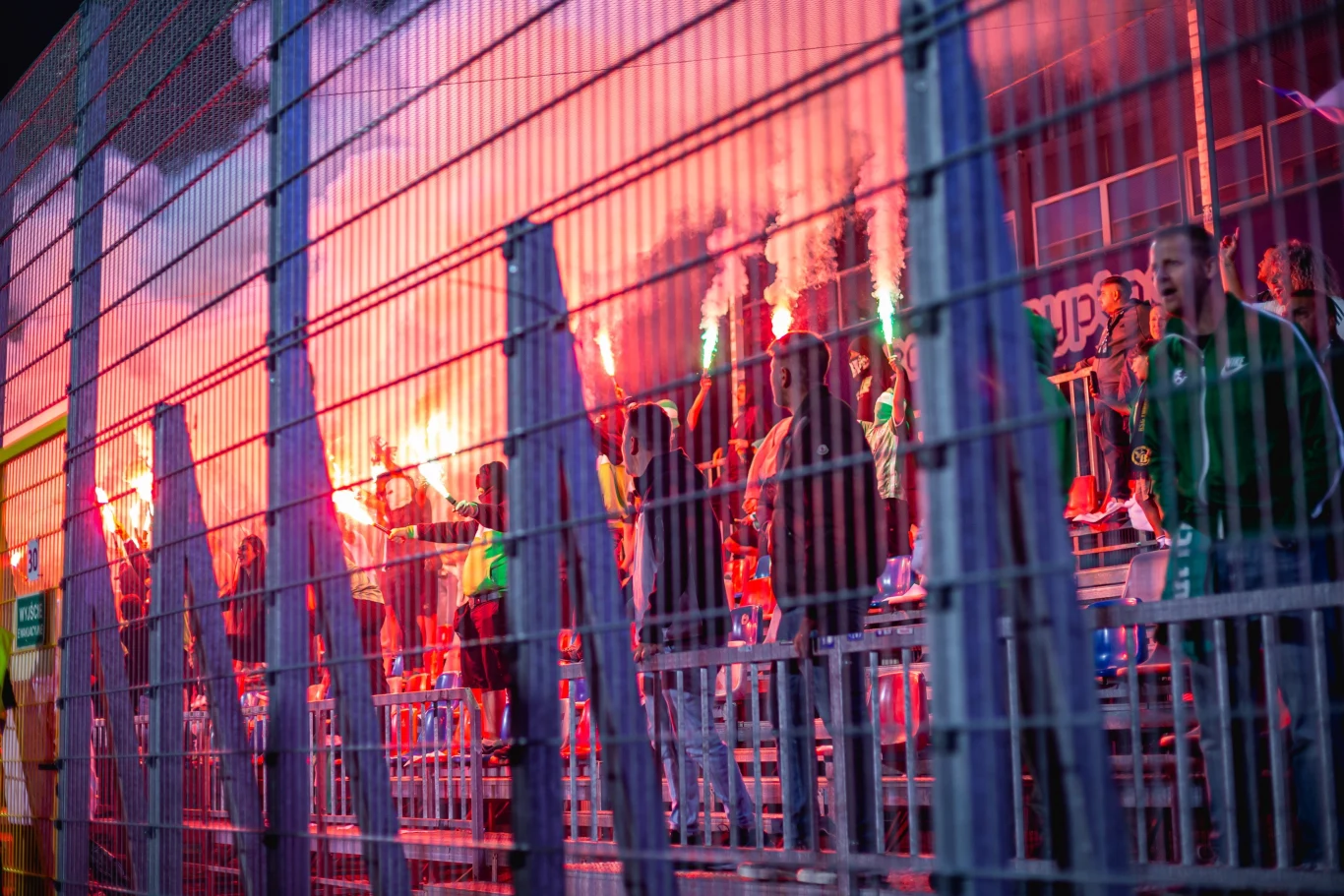 Kibice Maccabi Hajfa na stadionie Rakowa w Częstochowie Grupa kibiców na trybunach stadionu odpalająca race świetlne, intensywne czerwone światło i dym unoszący się nad tłumem, metalowe ogrodzenie oddzielające kibiców od boiska.