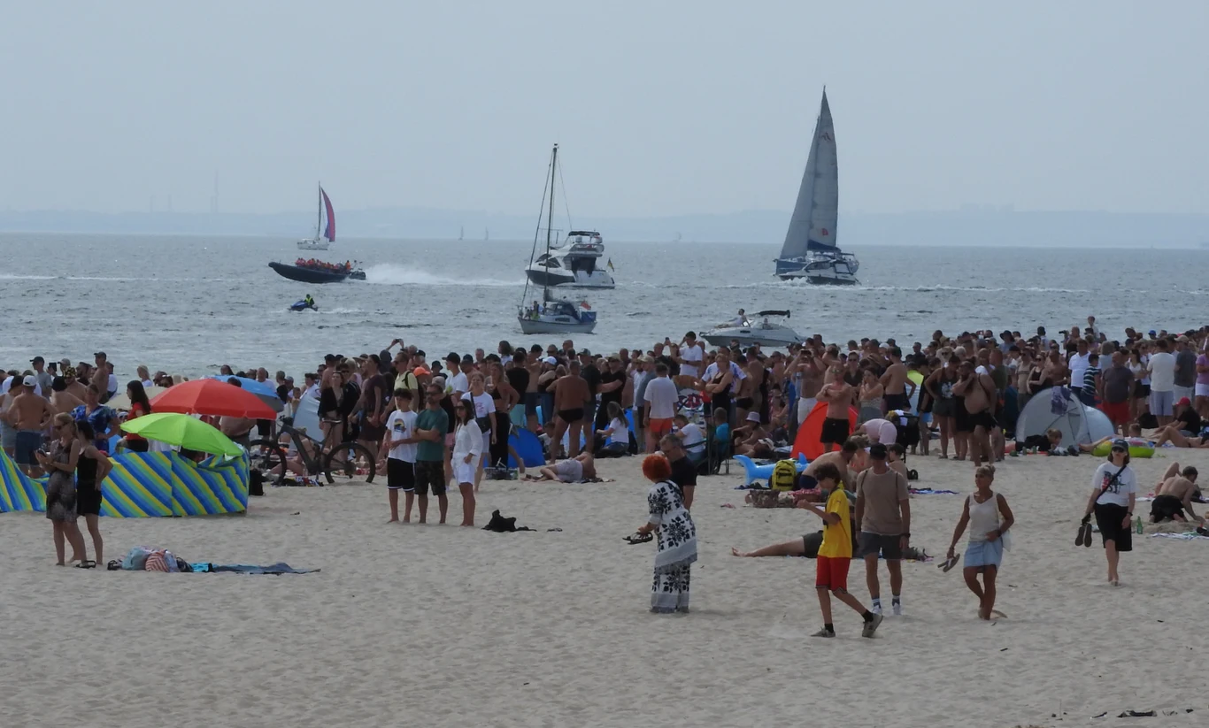 Tłum ludzi wypoczywających na piaszczystej plaży nad morzem, wiele osób opala się, dzieci bawią się piłką, w tle na wodzie żaglówki i motorówki, kolorowe namioty oraz parasole plażowe.