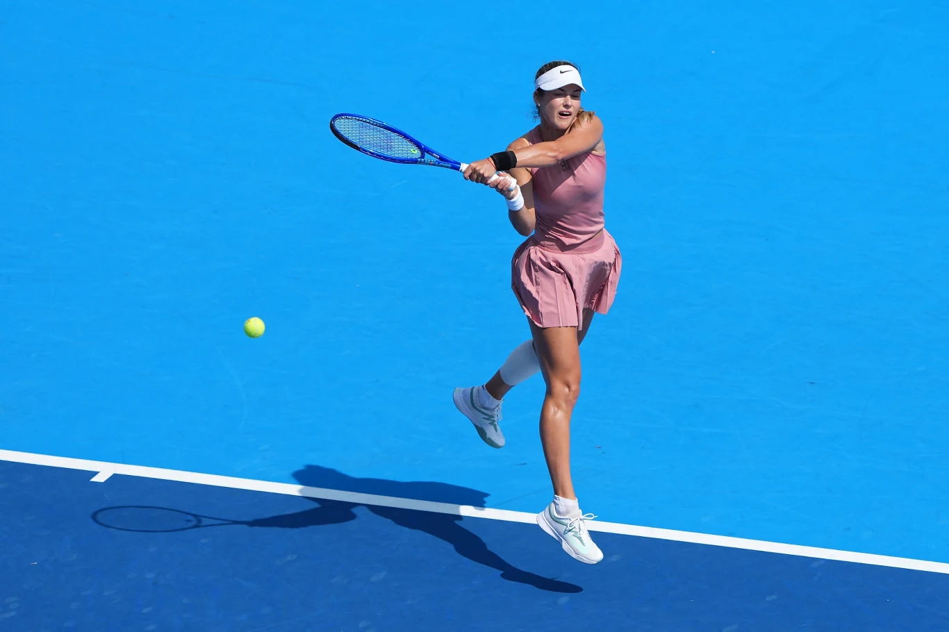 Tenisistka w różowej sukience wykonuje zamach rakietą podczas meczu na niebieskim korcie, skupiona na żółtej piłce tenisowej przelatującej obok niej.