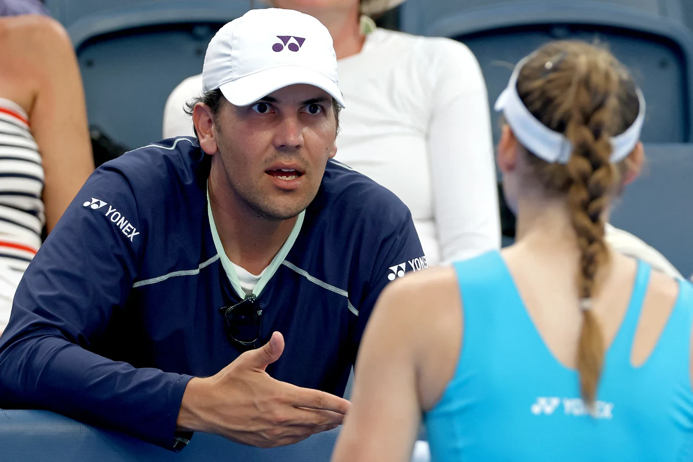 Mężczyzna w sportowej czapce i bluzie Yonex gestykuluje podczas rozmowy z tenisistką ubrana w niebieski strój sportowy, siedzącą przodem do niego na trybunach.