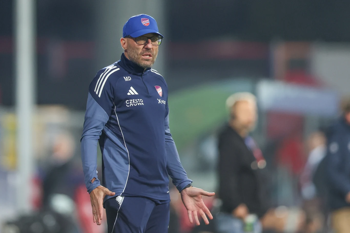 Marek Papszun Trener w sportowym ubraniu z logo klubu, stojący na boisku podczas meczu, w tle niewyraźne sylwetki innych osób.