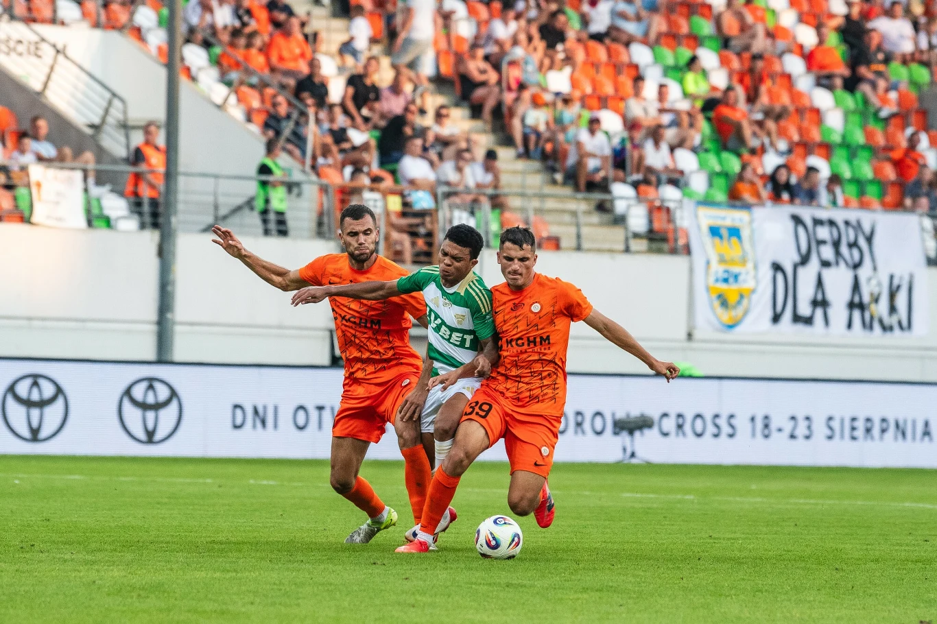 Dwóch piłkarzy w pomarańczowych strojach próbuje odebrać piłkę zawodnikowi w zielono-białym stroju na murawie stadionu, w tle trybuny z kibicami oraz transparenty.
