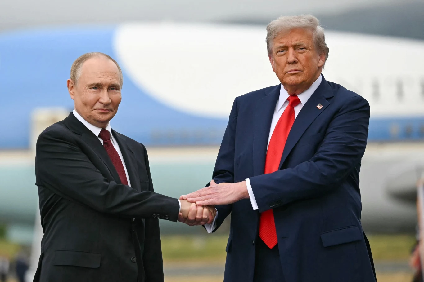 Amerykańskie media pisza o spotkaniu Trumpa z Putinem Dwóch mężczyzn w garniturach ściska sobie dłonie, w tle widać fragment samolotu.