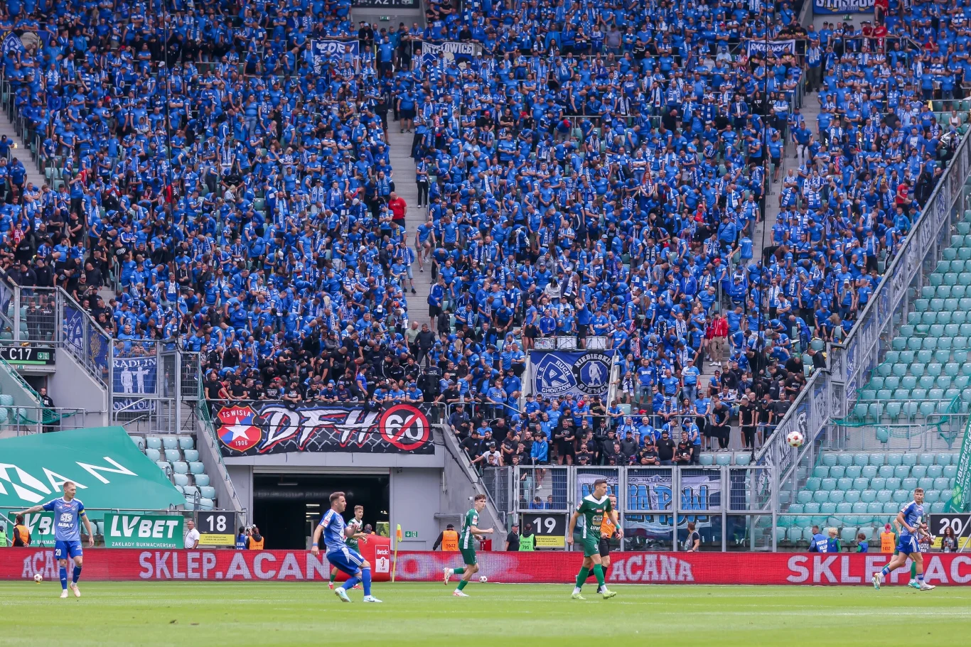 Kibice Ruchu Chorzów zawsze wypełniają sektor gości Piłkarze rywalizujący na boisku stadionu, w tle trybuny wypełnione kibicami ubranymi w niebieskie barwy, flagi oraz transparenty klubowe, energetyczna atmosfera sportowego wydarzenia.