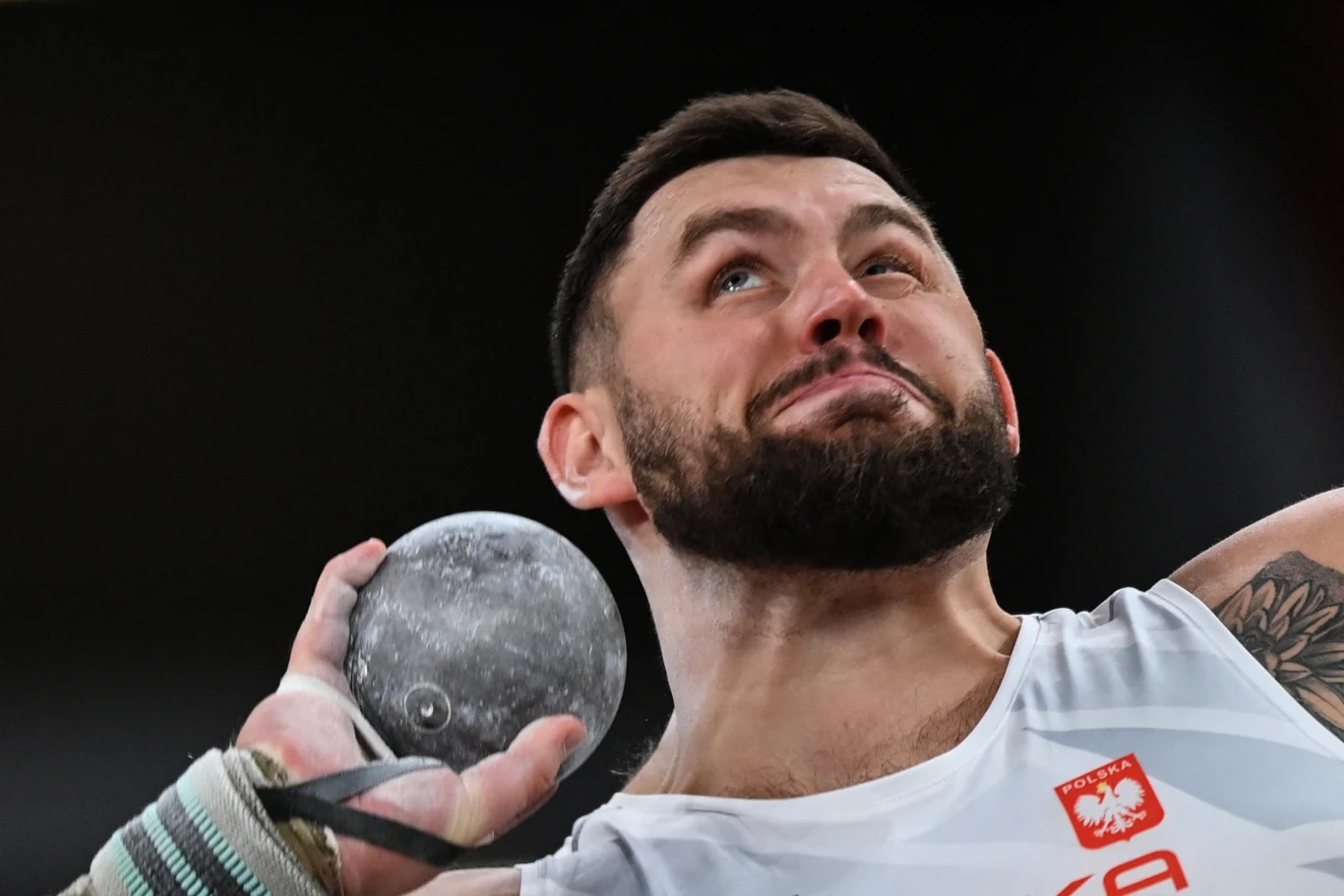 Sportowiec w trakcie pchnięcia kulą, skupiony wyraz twarzy, widoczny strój z polskim godłem oraz kuli w dłoni.