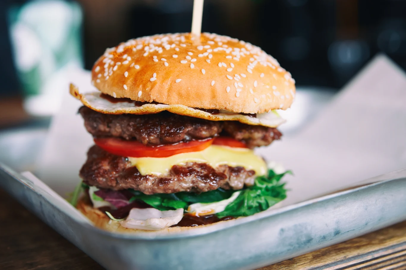 Dwuwarstwowy burger z wołowiną, plasterkami pomidora, liśćmi sałaty, żółtym serem, cebulą i sadzonym jajkiem w bułce z sezamem, umieszczony na tacy wyłożonej papierem.