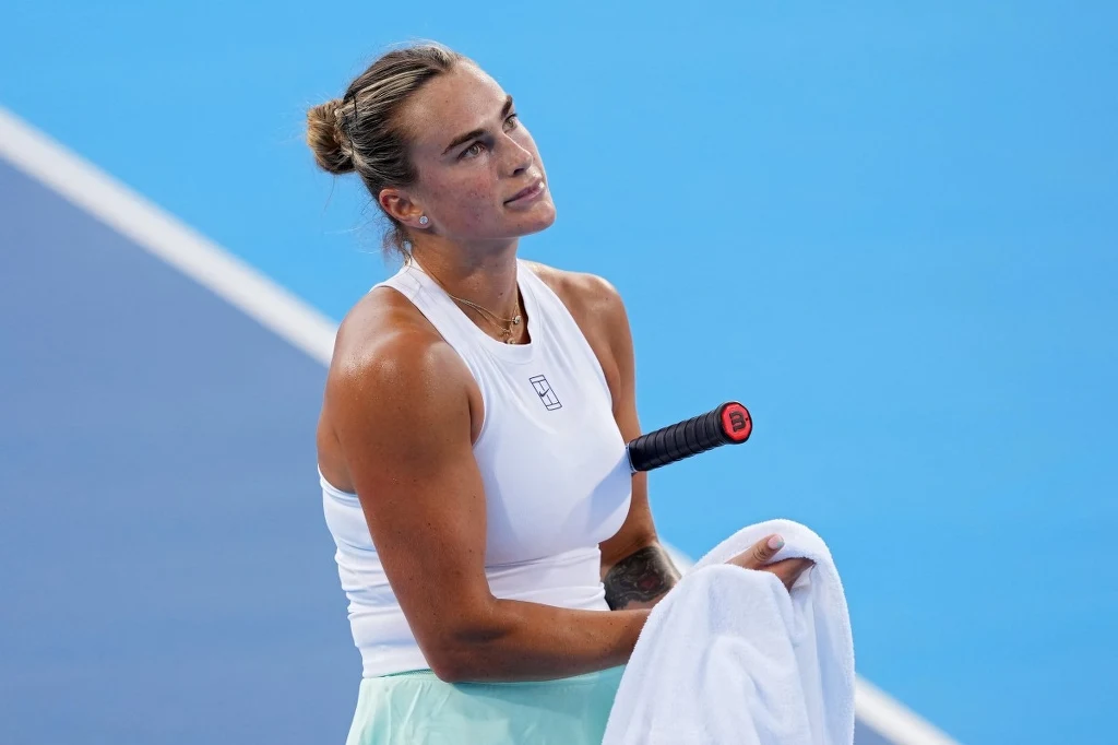 Aryna Sabalenka Zawodowa tenisistka podczas meczu na korcie trzymająca ręcznik, z rakietą tenisową wsuniętą w spodnie sportowe, skupiona i wyraźnie zamyślona.
