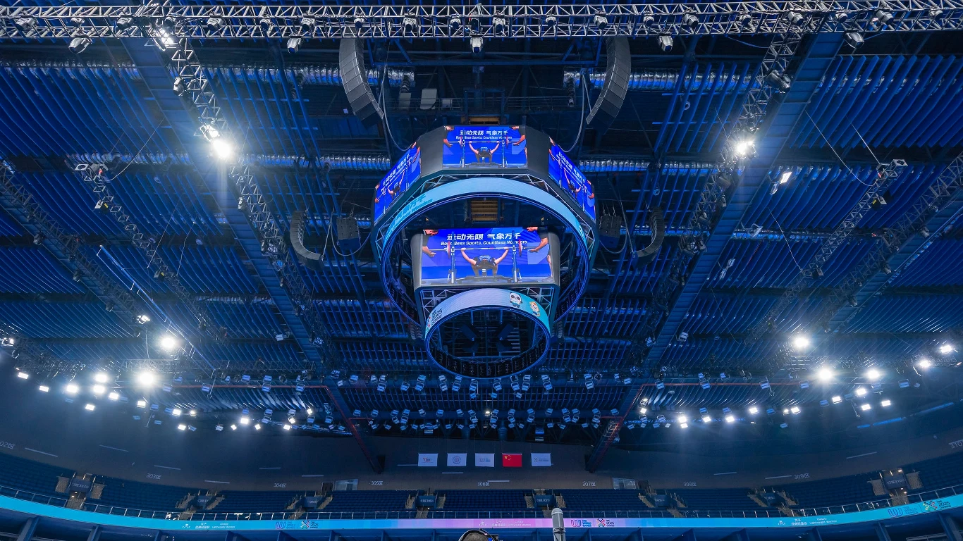Gigantyczny ekran LED zawieszony pod sufitem nowoczesnej hali sportowej, jasne oświetlenie sufitowe oraz stalowa konstrukcja wsporcza, trybuny w tle częściowo puste.