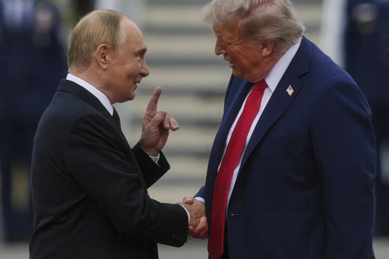 Władimir Putin i Donald Trump w Anchorage Dwóch mężczyzn ubranych w garnitury wymienia uścisk dłoni i patrzy na siebie z uśmiechami, jeden z nich unosi palec wskazujący w geście podkreślającym komunikację, w tle widoczne postacie w mundurach.
