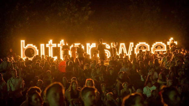 Tłum ludzi siedzący i stojący przed dużym, podświetlonym napisem "bittersweet", ciemne tło z wyraźnie widocznym światłem bijącym z napisu, klimat koncertu lub festiwalu.