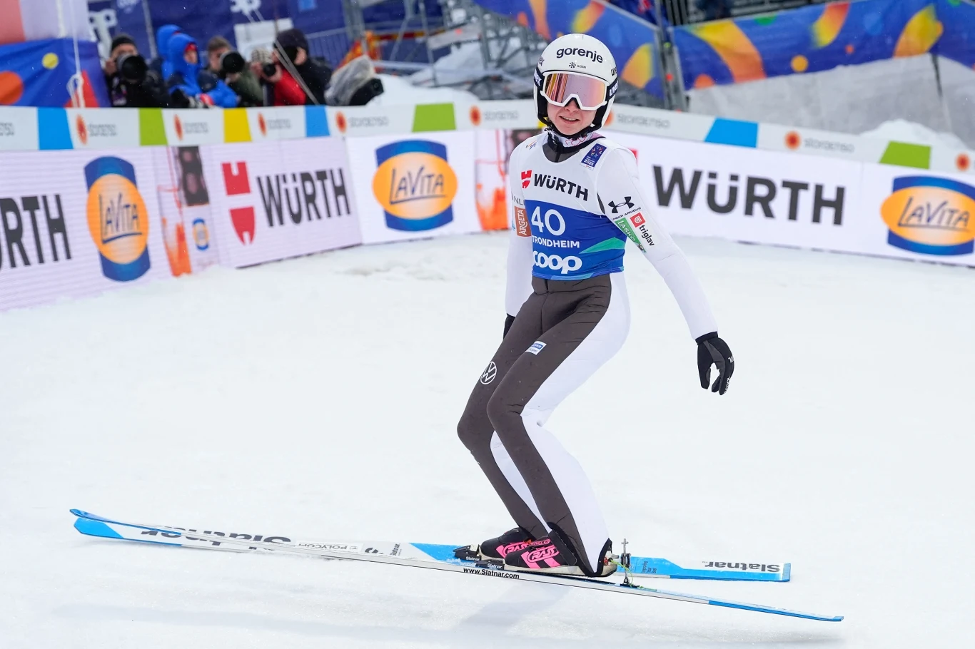 Zawodnik w kombinezonie narciarskim podczas lądowania po skoku na nartach na skoczni pokrytej śniegiem, w tle widoczne są bandy reklamowe oraz kibice.