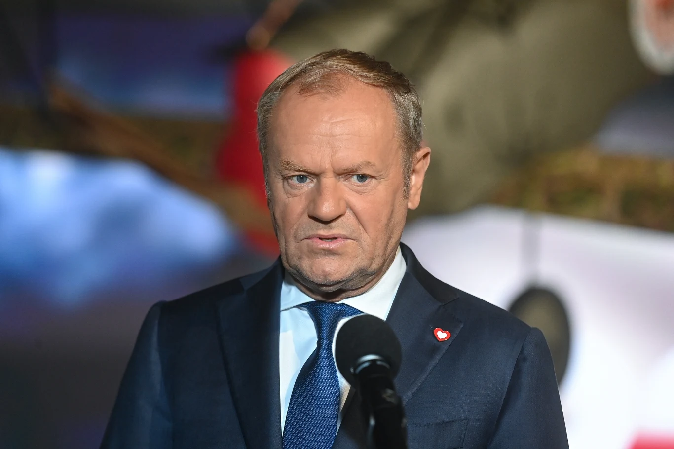 Donald Tusk Mężczyzna w średnim wieku w garniturze i niebieskim krawacie stoi przed mikrofonem, w tle niewyraźne elementy dekoracyjne oraz światła.