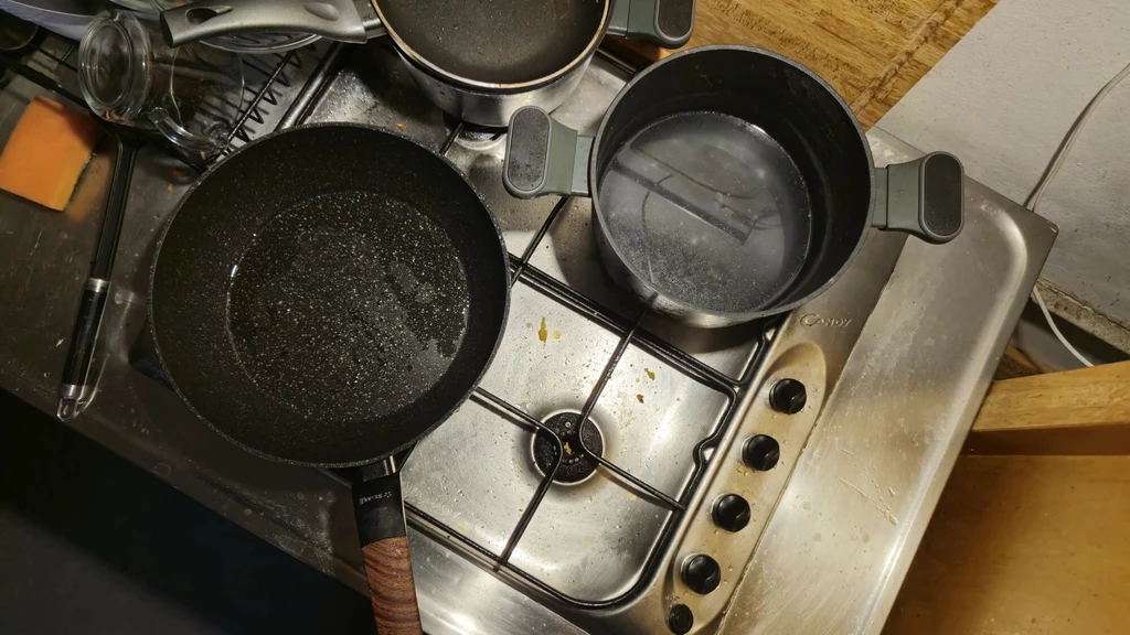Widok z góry na stalową kuchenkę gazową z dwoma garnkami i patelnią, obok widać naczynia oraz gąbkę do mycia. Naczynia stoją na palnikach, na powierzchni kuchenki widoczne są drobne resztki jedzenia i ślady użytkowania.