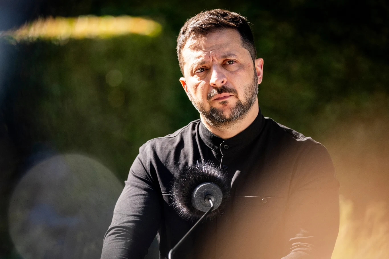 Wołodymyr Zełenski Mężczyzna o poważnym wyrazie twarzy stoi przed mikrofonem, ubrany w czarną koszulę; wokół rozmyte światło słoneczne, w tle rozmyte zielone elementy sugerujące otoczenie zewnętrzne.