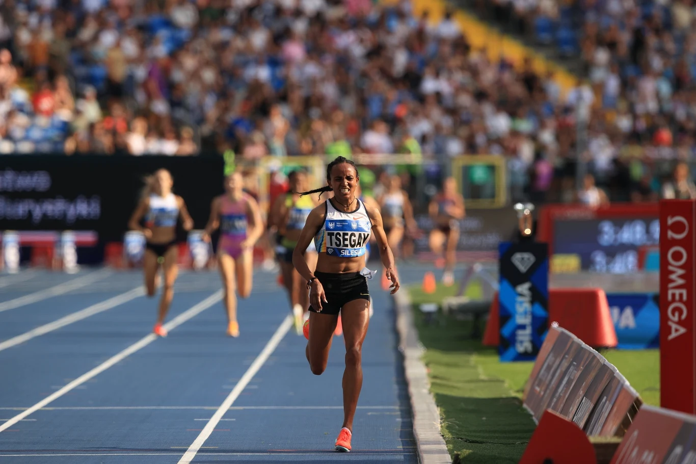 Zawodniczka biegnąca na czele na ostatniej prostej bieżni stadionu lekkoatletycznego, z widoczną tablicą wyników oraz publicznością w tle, pozostałe biegaczki za nią kończą wyścig.