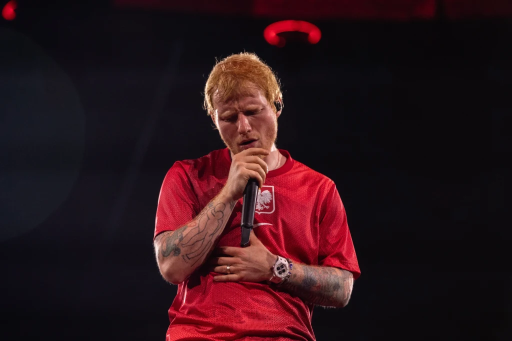 Ed Sheeran wystąpił na Tarczyński Arena we Wrocławiu Mężczyzna z rudymi włosami ubrany w czerwoną koszulkę z godłem Polski śpiewa do mikrofonu, trzymając jedną rękę na brzuchu i stojąc na ciemnym tle, oświetlony światłem scenicznym.