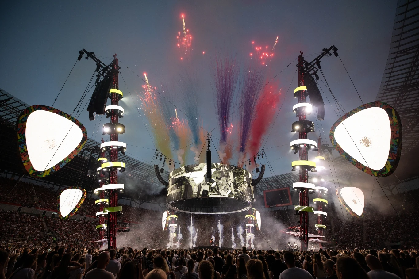 Tłum ludzi zgromadzony na dużym stadionie podczas widowiskowego koncertu. Wielka scena wyposażona w gigantyczne ekrany i reflektory, nad którymi wystrzeliwane są kolorowe fajerwerki oraz dymy w różnych barwach. Oświetlenie i efekty specjalne podkreślaj...