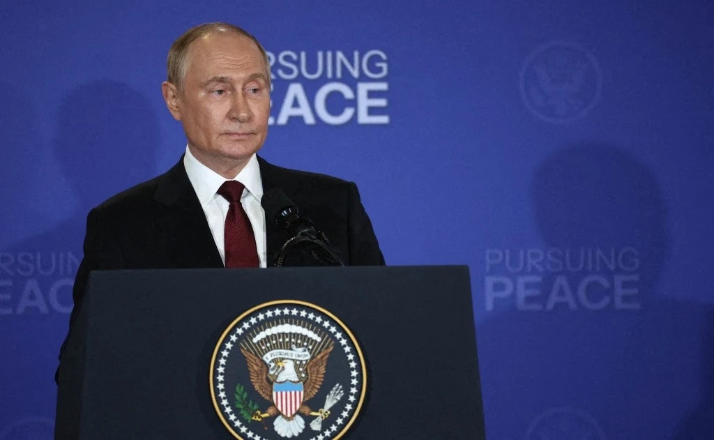 Rosyjski przywódca Władimir Putin po rozmowie z prezydentem USA Donaldem Trumpem Mężczyzna w garniturze stojący przy pulpicie z amerykańskim godłem prezydenckim na tle niebieskiej ściany z napisem 'Pursuing Peace'. Wyraz twarzy poważny, scena oficjalna.