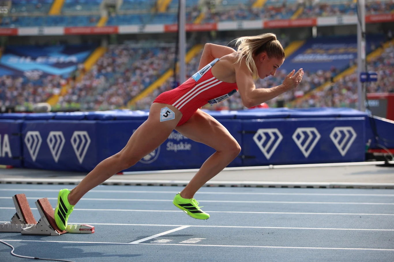 Natalia Bukowiecka Lekkoatletka w dynamicznym momencie startu biegu, ubrana w czerwony strój sportowy i jasnozielone buty, na tle stadionu z widownią.