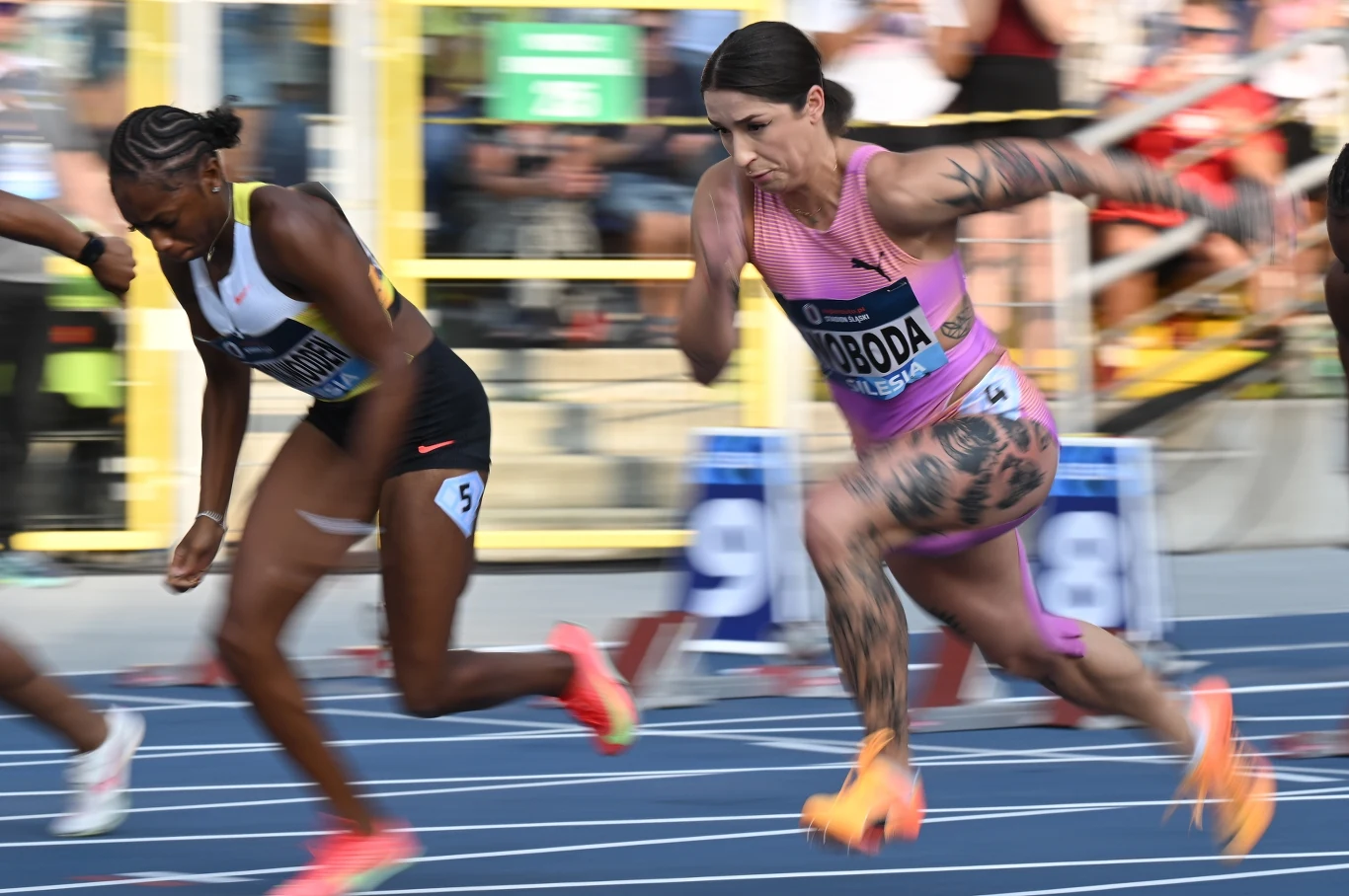 Dwie zawodniczki biegną na bieżni w trakcie wyścigu lekkoatletycznego, skupienie i determinacja widoczne na ich twarzach, dynamiczne ruchy i wyraźnie zaznaczone mięśnie podkreślają sportową rywalizację.