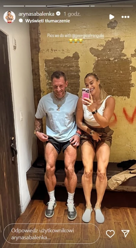 Aryna Sabalenka i Georgios Frangulis Dwie osoby siedzące na drewnianej ławce w prostym, surowym wnętrzu z odsłoniętą ceglaną ścianą, jedna z nich robi selfie smartfonem, obie uśmiechnięte i w sportowych ubraniach.