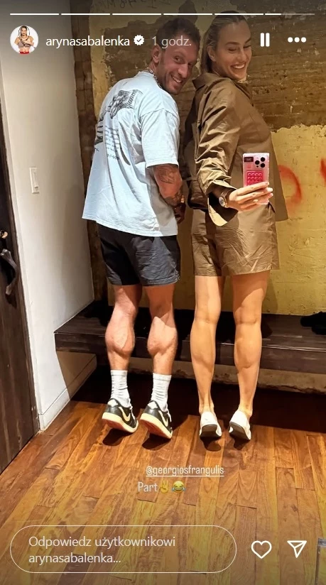 Aryna Sabalenka i Georgios Frangulis Uśmiechnięta kobieta w jasnobrązowej sukience i mężczyzna w jasnej koszulce oraz czarnych spodenkach pozują razem przed lustrem, kobieta trzyma telefon, na drewnianej podłodze i tle ceglanej ściany.