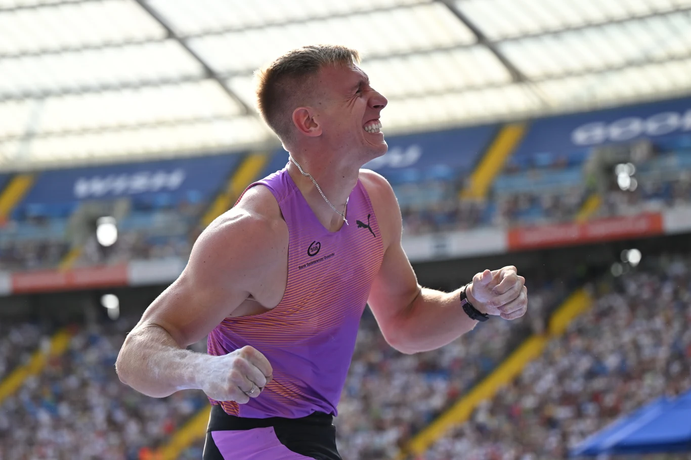 Sportowiec w stroju lekkoatletycznym z uniesionymi zaciśniętymi pięściami i wyrazem wysiłku na twarzy na tle stadionu pełnego publiczności.