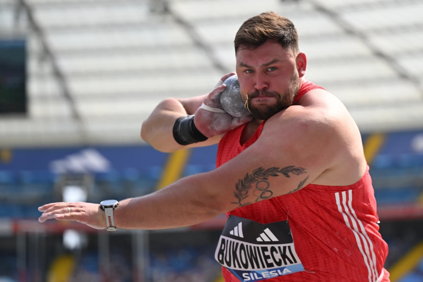 Konrad Bukowiecki Zawodnik w czerwonym stroju podczas zawodów pchnięcia kulą, skoncentrowany na wykonywaniu techniki, w tle widoczne trybuny stadionu.