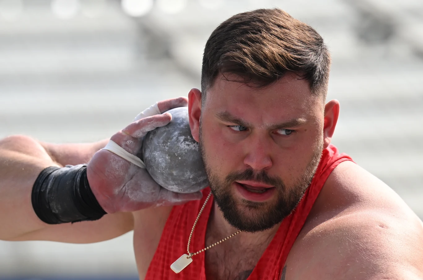 Konrad Bukowiecki Sportowiec w czerwonym stroju przygotowuje się do pchnięcia kulą, skupiony wyraz twarzy podkreśla koncentrację, dłoń objęta magnezją trzyma kulę przy szyi, w tle rozmyte trybuny stadionowe