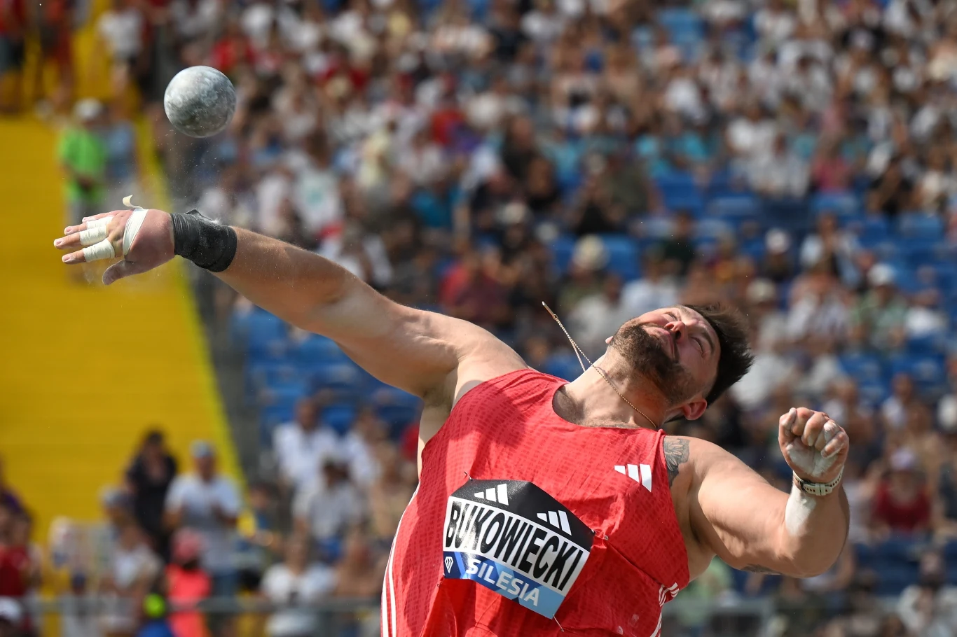 Konrad Bukowiecki Lekkoatleta w trakcie dynamicznego pchnięcia kulą podczas zawodów lekkoatletycznych, z widoczną kulą w powietrzu oraz tłumem kibiców na trybunach w tle.