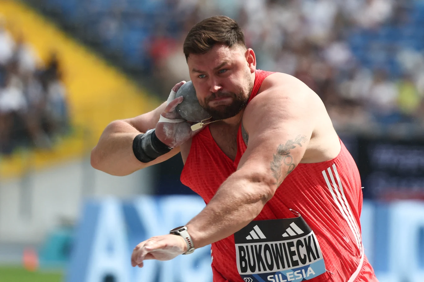 Konrad Bukowiecki Postawny mężczyzna w czerwonym stroju sportowym podczas pchnięcia kulą na stadionie, skupiona twarz i wyraźnie napięte mięśnie, w tle rozmyta publiczność oraz bieżnia.