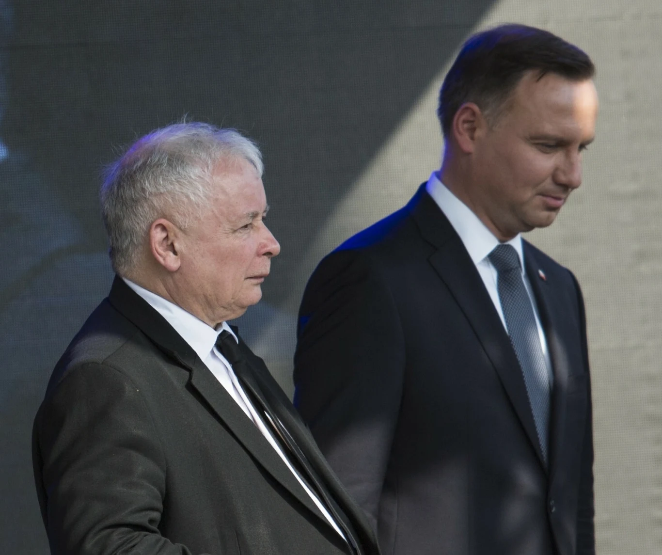 Prezes PiS Jarosław Kaczyński i były prezydent Andrzej Duda Dwóch mężczyzn w garniturach na tle jasnej ściany, ustawionych bokiem do obiektywu, jeden z nich patrzy poważnie przed siebie, drugi lekko spuszcza wzrok.