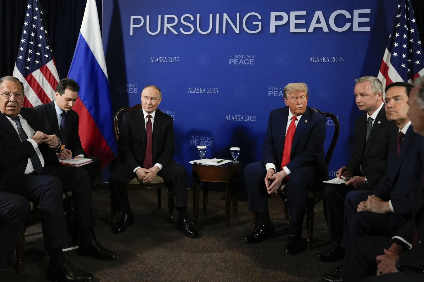Media informują o wycieku tajnych dokumentów po szczycie Trump-Putin Grupa polityków siedzących naprzeciwko siebie podczas oficjalnego spotkania, po lewej stronie widoczna flaga Rosji, po prawej flagi Stanów Zjednoczonych, na tle niebieskiego baneru z napisem 'Pursuing Peace'.