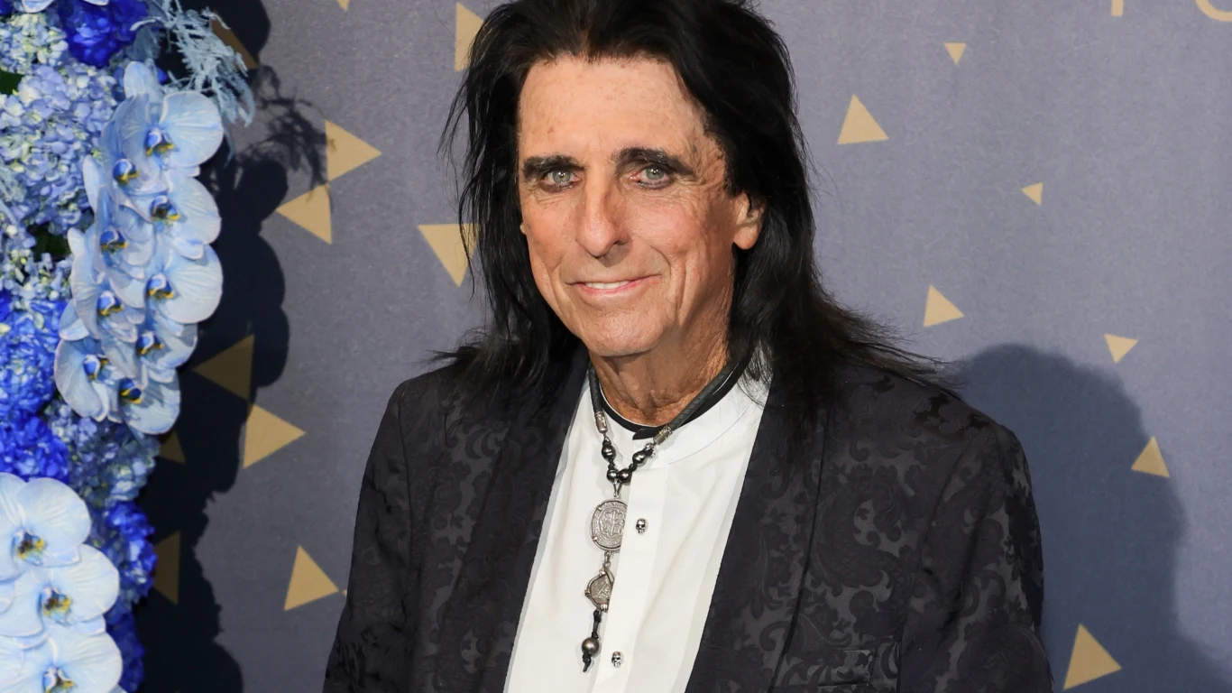 Alice Cooper Mężczyzna z długimi ciemnymi włosami ubrany w elegancką marynarkę o ozdobnym wzorze i białą koszulę, stojący przed dekoracją z niebieskich i fioletowych kwiatów oraz geometrycznych wzorów na ścianie.