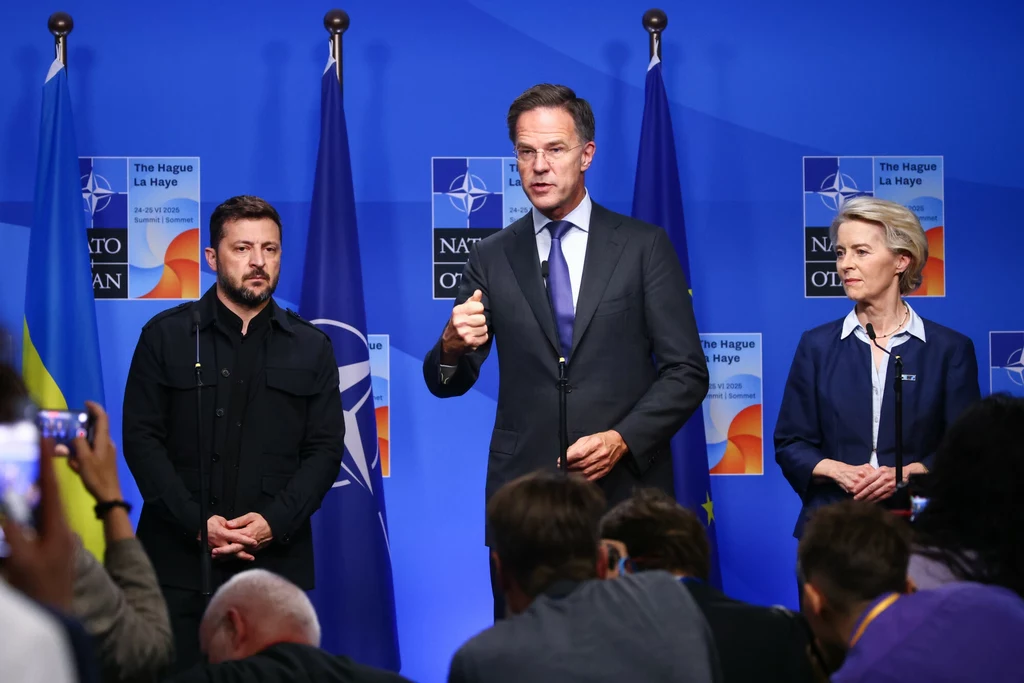 Trzech polityków stojących przed mikrofonami na tle flag NATO, Ukrainy i Unii Europejskiej, środkowy mężczyzna gestykuluje podczas przemówienia, po bokach stoją pozostali uczestnicy konferencji, poniżej widać zgromadzonych dziennikarzy.