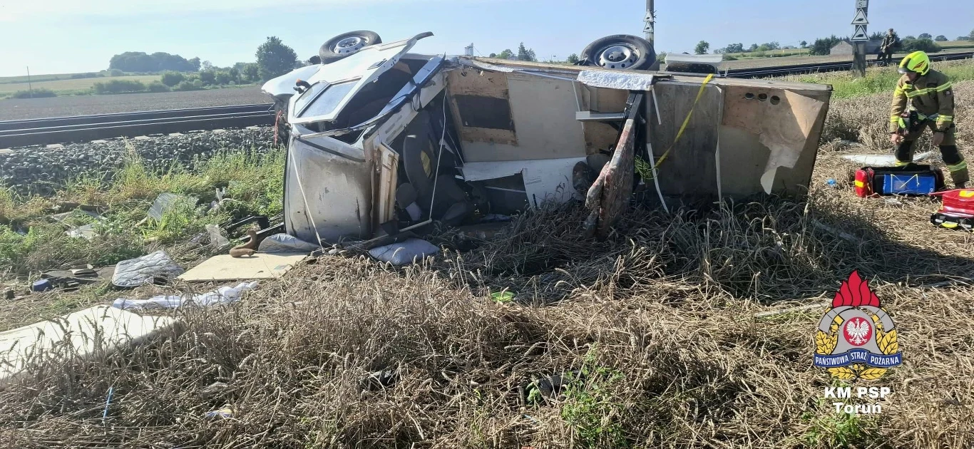 Rozbity samochód dostawczy leży na boku w pobliżu torów kolejowych, wokół rozrzucone są elementy pojazdu i rzeczy, z prawej strony widoczny strażak w żółtym mundurze świadczący o prowadzeniu akcji ratunkowej.