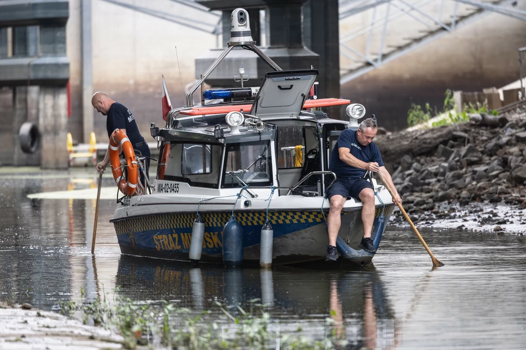 Instytut Meteorologii i Gospodarki Wodnej wydał 31 ostrzeżeń o suszy hydrologicznej Łódź patrolowa Straży Miejskiej płynąca przy brzegu rzeki, na pokładzie dwóch funkcjonariuszy zbierających coś z powierzchni wody długimi narzędziami, w tle betonowe konstrukcje mostu i przybrzeżne rośliny.