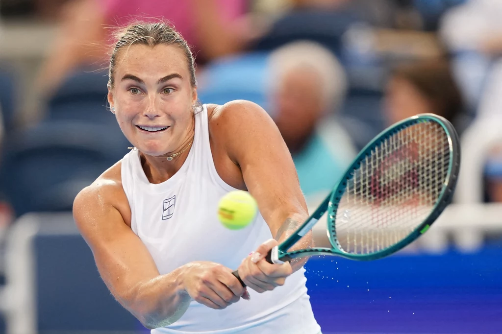 Aryna Sabalenka Zawodniczka w białej koszulce skupiona podczas odbicia piłki tenisowej rakietą, w tle rozmazani widzowie na stadionie.