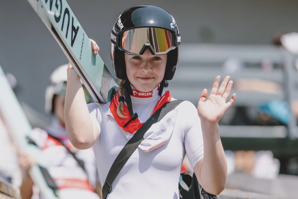 Pola Bełtowska Skoczkini narciarska w białym kombinezonie i kasku z goglami, trzymająca narty na ramieniu oraz machająca ręką w kierunku aparatu, wokół niej rozmyte sylwetki innych osób oraz elementy trybun.