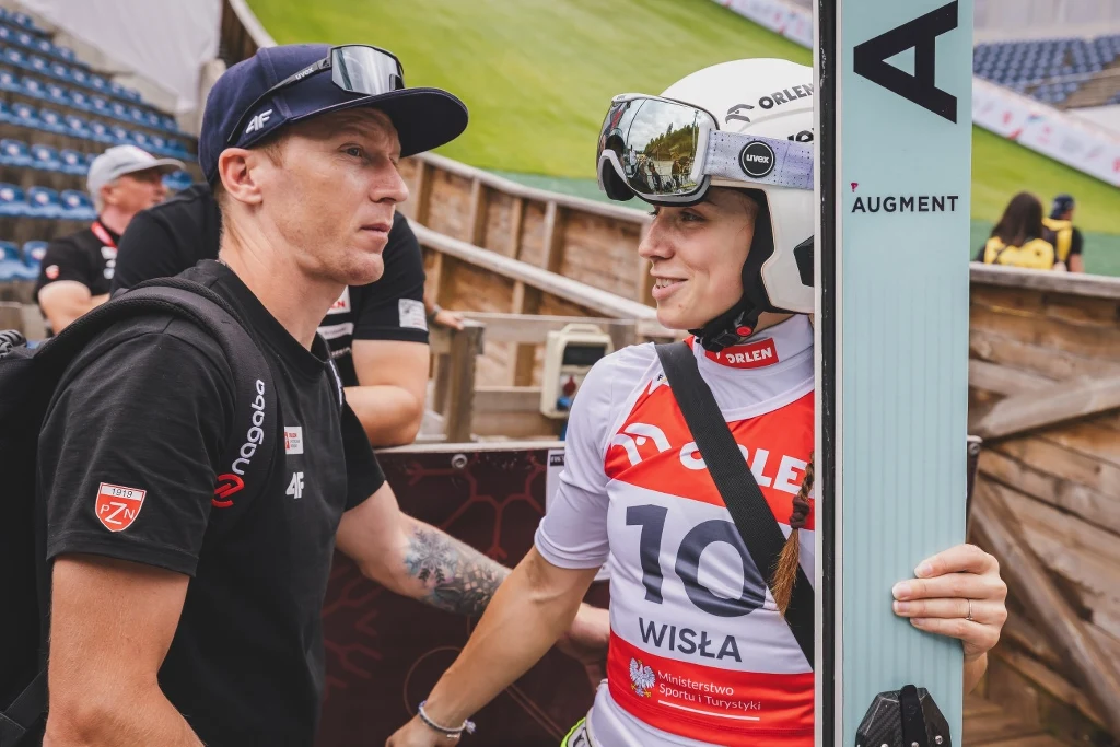 Dwójka sportowców w otoczeniu skoczni narciarskiej, jedna osoba ubrana w strój skoczka narciarskiego z numerem startowym 10, trzymająca narty i rozmawiająca z trenerem lub członkiem obsługi.