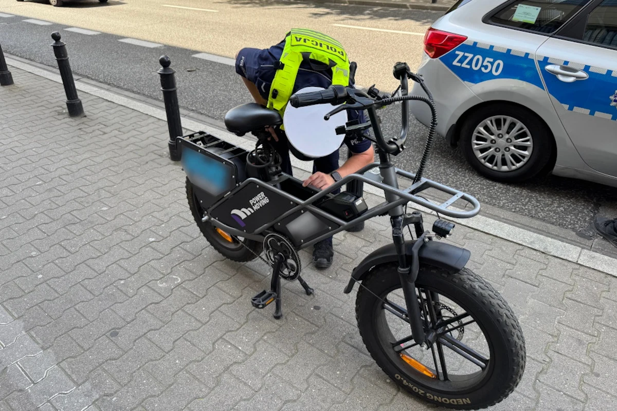Policjant sprawdza elektryczny rower na chodniku przy ulicy, w tle zaparkowany radiowóz policyjny, a wszystko dzieje się w miejskim otoczeniu.