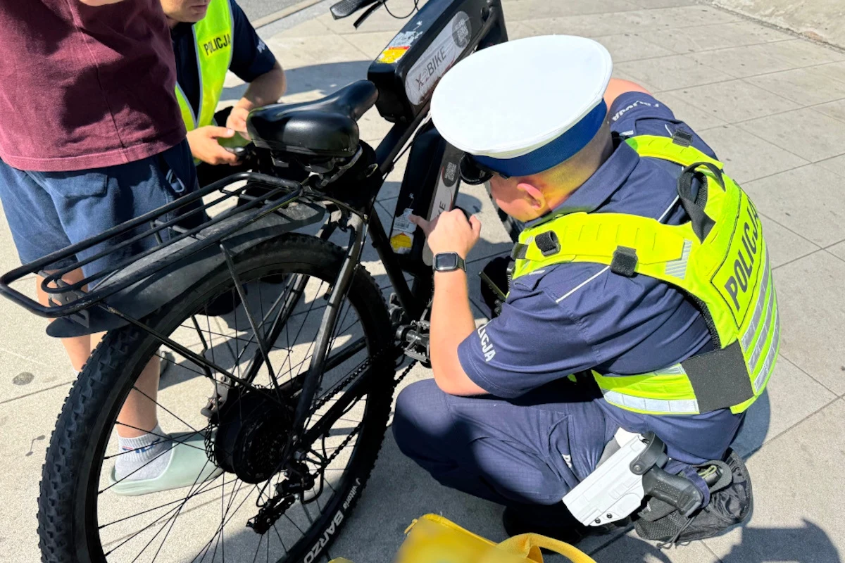 Policjant w żółtej kamizelce odblaskowej sprawdza rower elektryczny stojący na chodniku, obok stoi dziecko ubrane w krótkie spodenki i bordową koszulkę.
