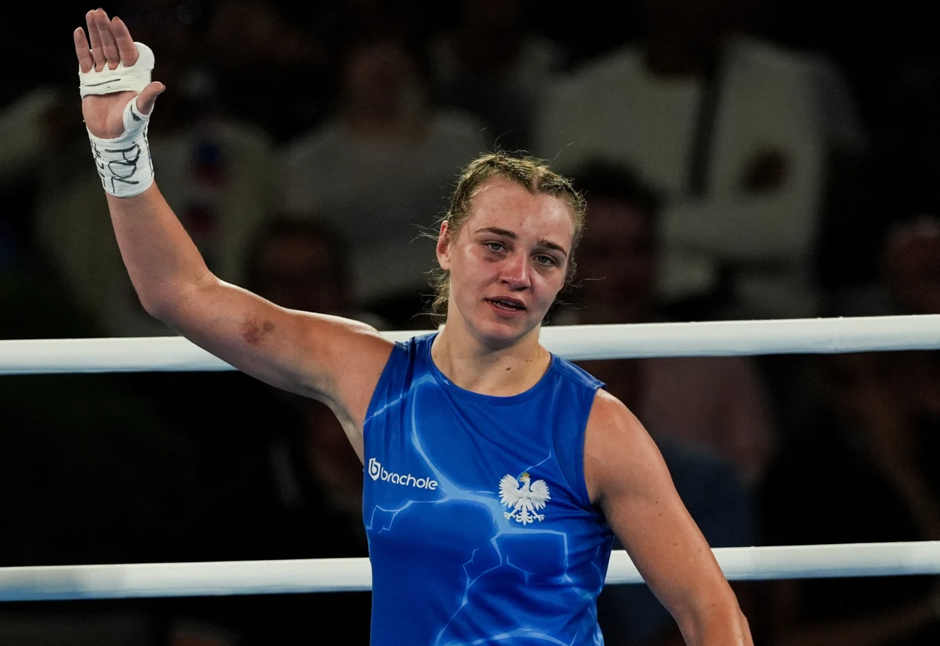 Julia Szeremeta Bokserka w niebieskim stroju z polskim godłem na piersi podnosi rękę w geście zwycięstwa, na twarzy widoczne emocje i ślady zmęczenia, w tle rozmyte sylwetki widzów.
