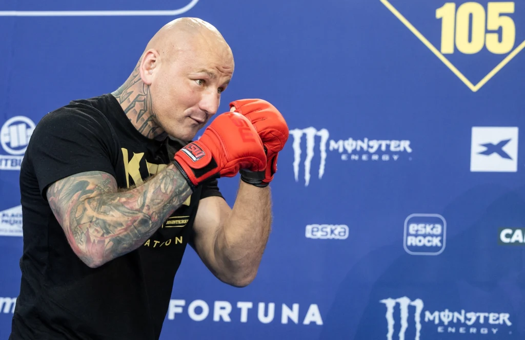 silnie zbudowany mężczyzna w czerwonych rękawicach bokserskich przyjmuje postawę do walki na tle niebieskiej ściany z logotypami sponsorów