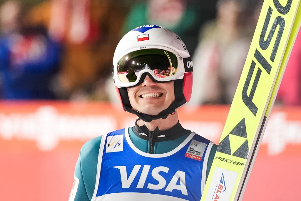 Kamil Stoch Uśmiechnięty skoczek narciarski w kasku i goglach, ubrany w kombinezon sportowy z widocznym logo sponsora, trzyma narty w otoczeniu rozmytej publiczności.