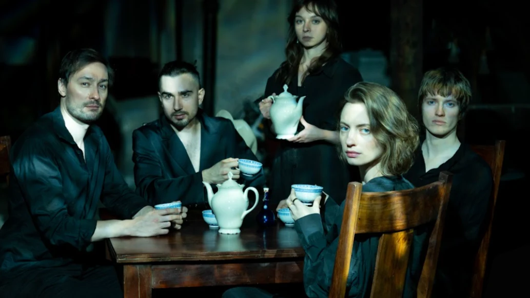 noco Pięć osób siedzi przy drewnianym stole, wszyscy ubrani w ciemne ubrania, skupieni wokół filiżanek i imbryków, wpatrując się poważnie w stronę obiektywu, w tle rozmyte cienie.