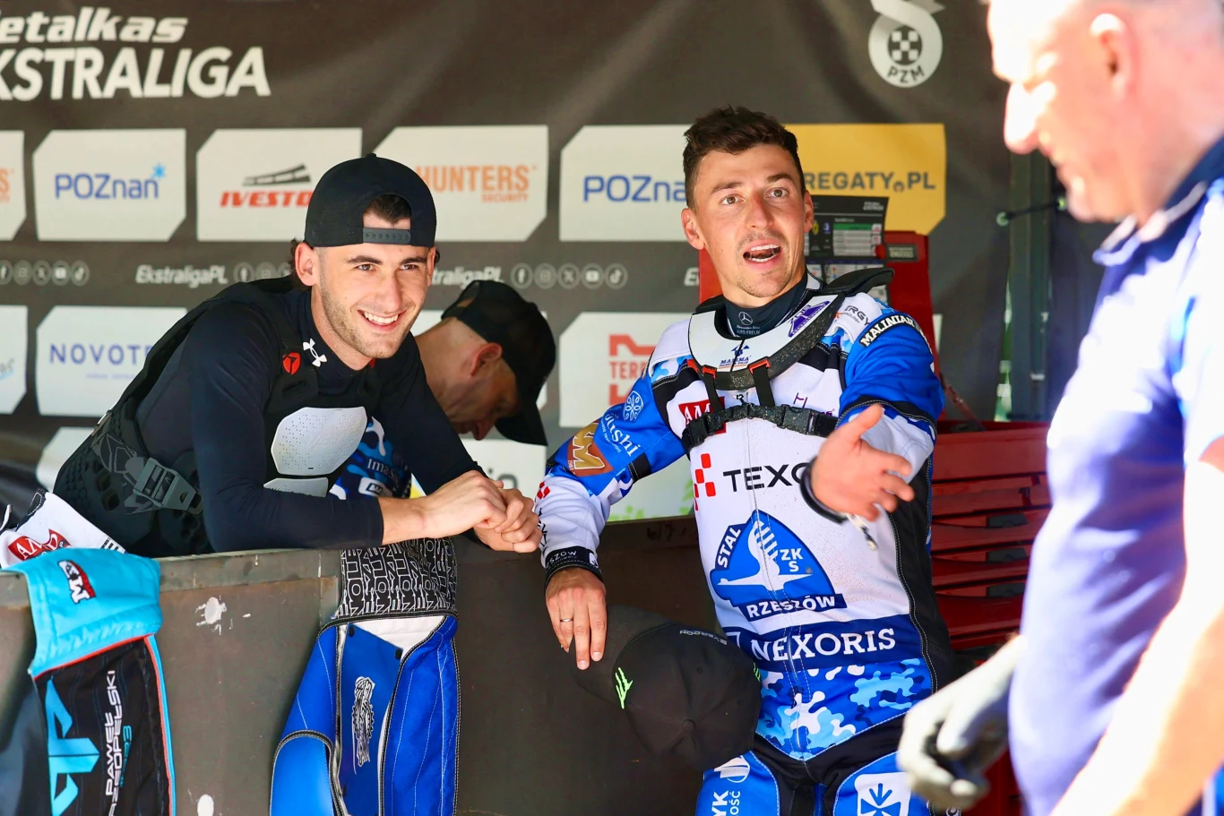 Dwóch żużlowców w kombinezonach zespołu TEXOM Stal Rzeszów rozmawia przy boksie serwisowym podczas zawodów, w tle widoczne banery sponsorów i fragment motocykla.